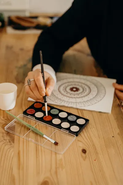 Close-up of hand painting a colorful mandala with watercolors on wooden table.