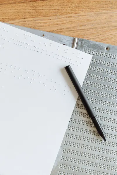 A vertical shot of Braille paper and a pen on a wooden table, highlighting tactile writing.