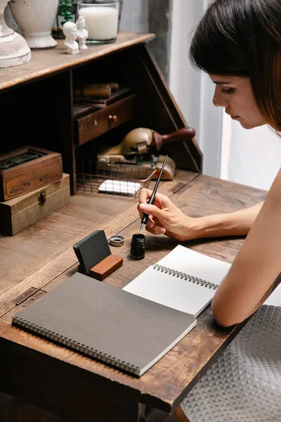 A woman writes in a notebook at a vintage wooden desk with ink and pen.