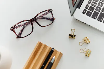 Top view of a stylish desk setup with eyeglasses, a laptop, paper clips, and pens on a white table.