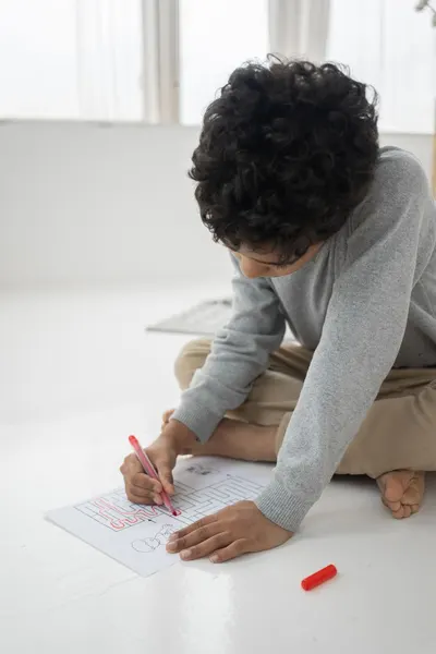 Concentrated anonymous African American boy with felt pen coloring maze on paper while sitting on floor in room at home