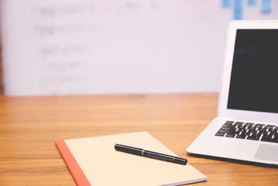 Modern workspace featuring a laptop, notebook, and pen on a wooden desk with a blurred whiteboard in the background.