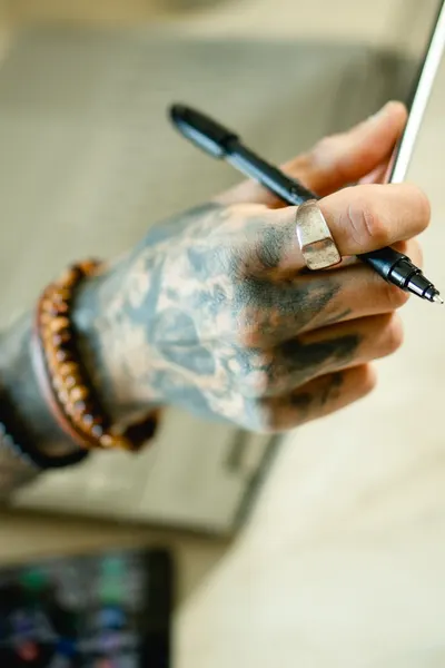 Close-up of a tattooed hand holding a pen, adorned with a ring, over a blurred background.