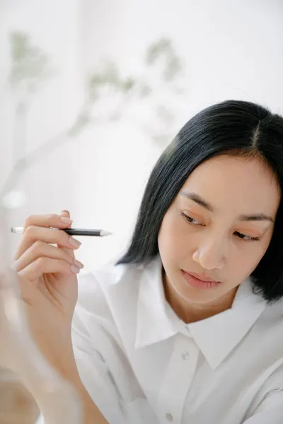 Pensive young Asian female in white shirt raising pen in hand while looking away behind blurred vase with twig
