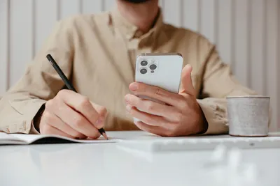 A man writes in a notebook while holding a smartphone at an office desk.