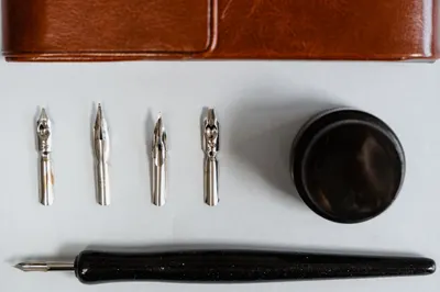 A flat lay of calligraphy nibs, holder, ink bottle, and leather case on a white background.