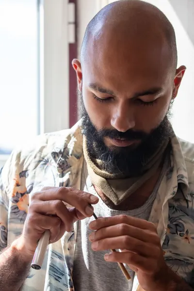Close-up of a bald, bearded man rolling a cannabis joint indoors with natural light.