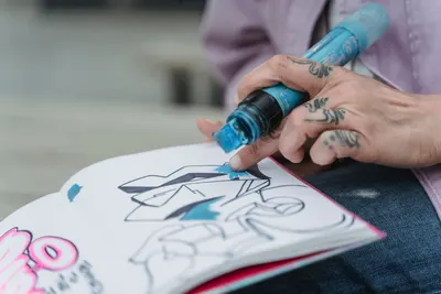 Crop anonymous artist with tattoos drawing graffiti in sketchbook while sitting on street on blurred background