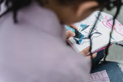 From above of crop unrecognizable female artist drawing sketches with marker in notebook while sitting in daylight