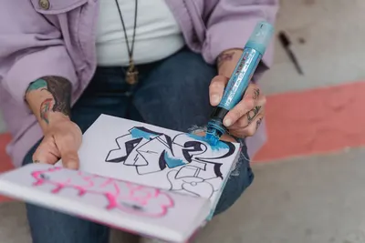 From above of crop anonymous female artist in denim ans with tattoos drawing on paper with marker while sitting in street