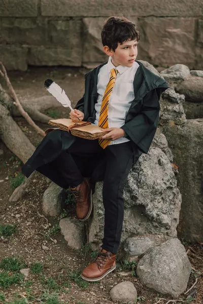 A child dressed as a wizard sitting outdoors with a quill and spell book, evoking a magical theme.