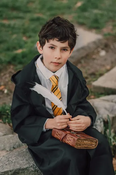 Child dressed as young wizard with a quill and spell book, seated outdoors.