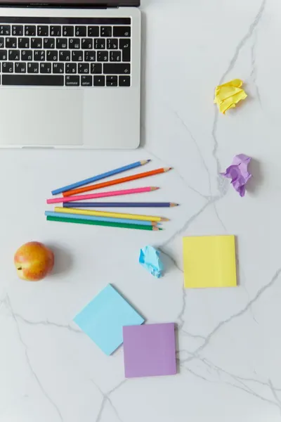 A top-down view of colored pencils, sticky notes, a laptop, and an apple on a white desk.