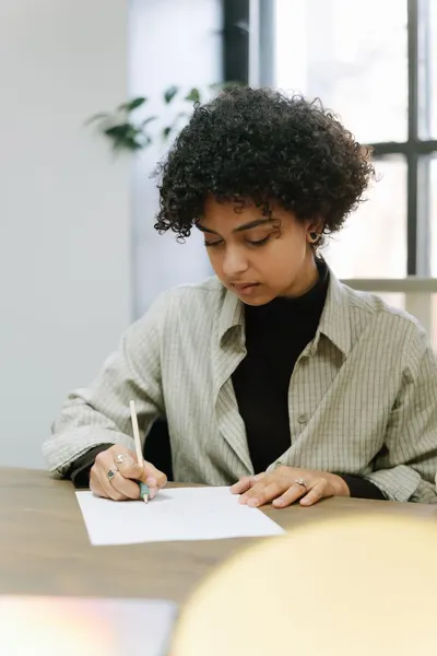 African American woman in a striped shirt writing on paper at a desk indoors.