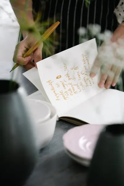 Close-up of hands writing a recipe with a pen in a cookbook at a dining table.