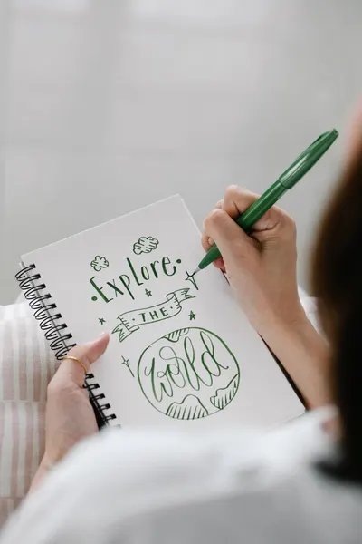 A person writing a motivational quote 'Explore the World' in green ink on a notebook.