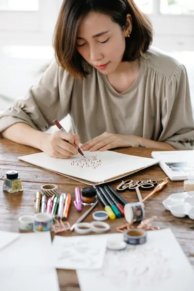 Female artist focused on creative hand lettering with assorted supplies on wooden desk.