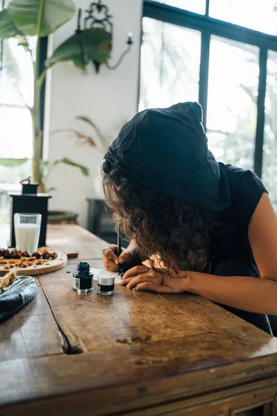 A woman with curly hair writing with an ink pen on a rustic wooden table indoors.