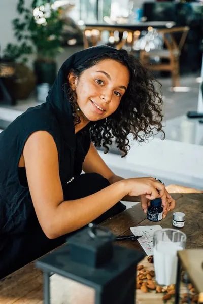 A cheerful woman with curly hair wearing a black hoodie holds a small ink bottle while sitting at a wooden table indoors.