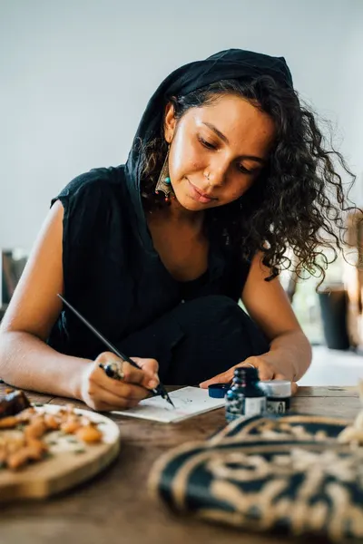 A focused woman writes creatively indoors, surrounded by ink and snacks on a wooden table.