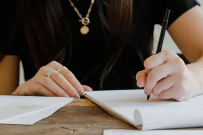 Close-up of a woman's hands writing in a notebook at a wooden desk, wearing jewelry.