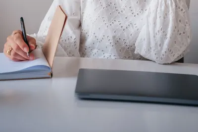 Close-up of a woman writing in a notebook while using a laptop at a desk.