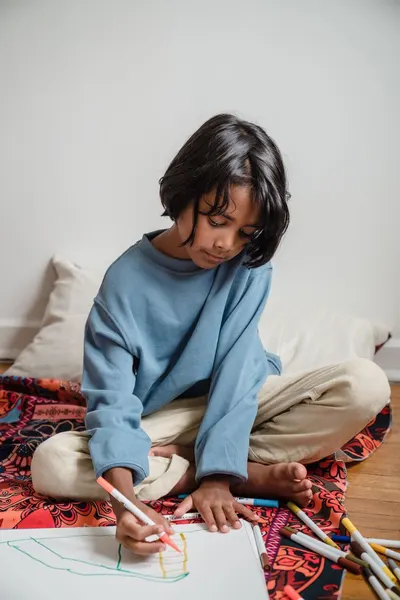 A young child drawing with felt tip pens, sitting cross-legged indoors.