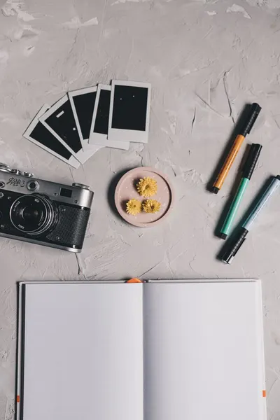 Top view of a creative workspace featuring a camera, open notebook, polaroids, and colorful markers on a textured background.