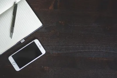 A clean and minimalist workspace featuring a smartphone, notebook, and pen on a dark wood table.