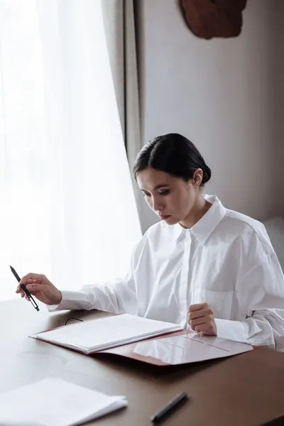 A woman in a white shirt diligently reviewing documents at a home office desk.