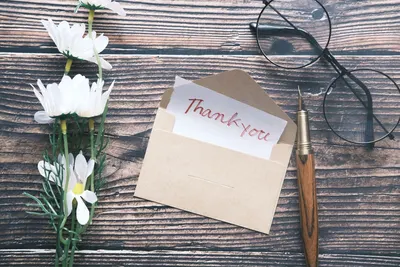 A flat lay of a thank you note in an envelope with daisies, glasses, and a pen on a wooden surface.